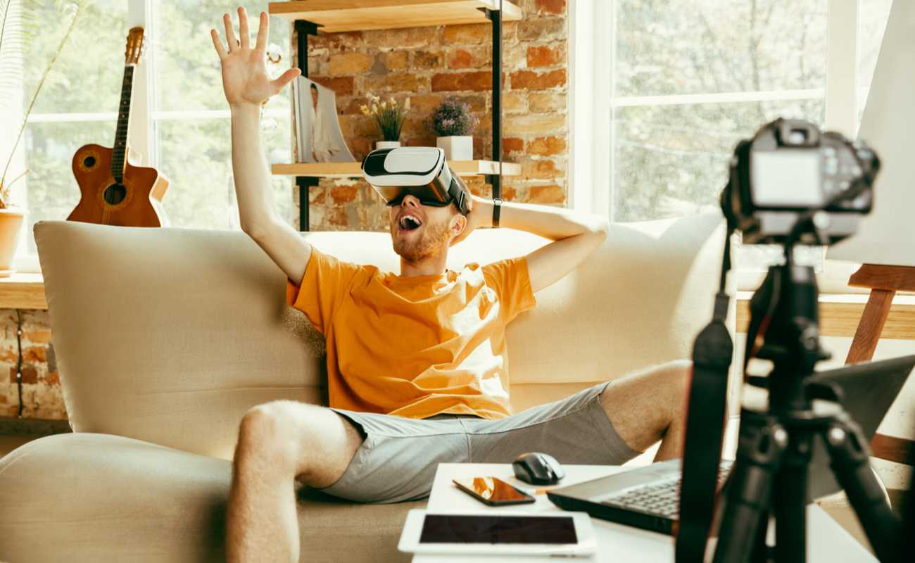 A man sitting on a coach with a VR headset while live streaming with a digital camera