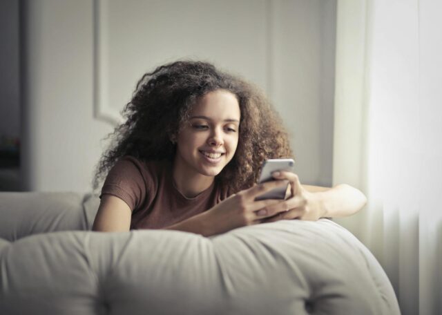 Photo of Smiling Woman Sitting on a Gray Bean Bag While Using Her Phone